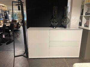 Modernes weißes Sideboard mit glatten Fronten, bestehend aus einer Tür und drei Schubladen. Obenauf stehen zwei hohe, bauchige Vasen aus dunklem Rauchglas. Links daneben steht eine außergewöhnliche schwarze Stehleuchte mit geometrischem Rahmen und einer sichtbaren Glühbirne. Im Hintergrund sind stilvoll eingerichtete Wohnbereiche mit Tischen, Stühlen und Regalen zu sehen.