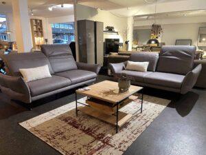 Zwei moderne, braune Ledersofas mit hohen Rückenlehnen und weißen Zierkissen stehen sich gegenüber auf einem beigefarbenen Teppich mit roten Akzenten. Dazwischen befindet sich ein rechteckiger Couchtisch mit Holzplatte und Metallgestell. Im Hintergrund ist ein stilvoll eingerichteter Möbel-Showroom mit weiteren Sitzgruppen und Accessoires zu sehen.