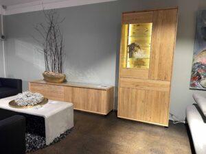 Moderne Wohnzimmereinrichtung mit einem Lowboard und einem beleuchteten Vitrinenschrank aus hellem Massivholz. Auf dem Lowboard steht eine große Schale mit dekorativen Zweigen, die Vitrine zeigt Glasböden mit stilvollen Deko-Objekten vor einer indirekt beleuchteten Rückwand. Die Möbel stehen vor einer hellgrauen Wand und werden von einem schlichten, rechteckigen Couchtisch in Betonoptik ergänzt.