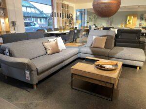 Zwei moderne graue Sofas mit unterschiedlichen Designs stehen in einer stilvollen Möbelausstellung einander gegenüber. Beide Sofas sind mit dekorativen Kissen ausgestattet. Dazwischen steht ein quadratischer Couchtisch mit Holzplatte und schwarzem Metallrahmen, darauf liegt eine Schale mit Holzdekoration. Im Hintergrund sind weitere Möbelstücke wie Tische, Stühle und Regale ausgestellt.