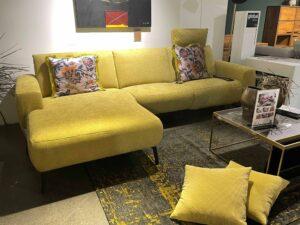 Modernes, senfgelbes Ecksofa mit floral gemusterten Zierkissen und verstellbarer Kopfstütze in einer stilvollen Ausstellung. Das Sofa steht auf einem dunklen Teppich mit gelben Akzenten. Vor dem Sofa befindet sich ein Glastisch mit goldenem Metallrahmen und dekorativen Elementen sowie zwei passende gelbe Kissen auf dem Boden.