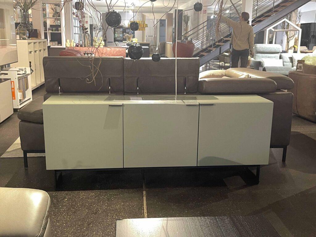 Modernes Sideboard in hellgrauer Hochglanzoptik steht vor einem braunen Ledersofa in einer stilvollen Möbelausstellung. Über dem Sideboard hängen dekorative schwarze Glaskugeln mit Pflanzenzweigen. Im Hintergrund sind weitere Möbelstücke wie Sofas, Sessel und Regale zu sehen, sowie ein Mann, der auf der Treppe Dekoration arrangiert.