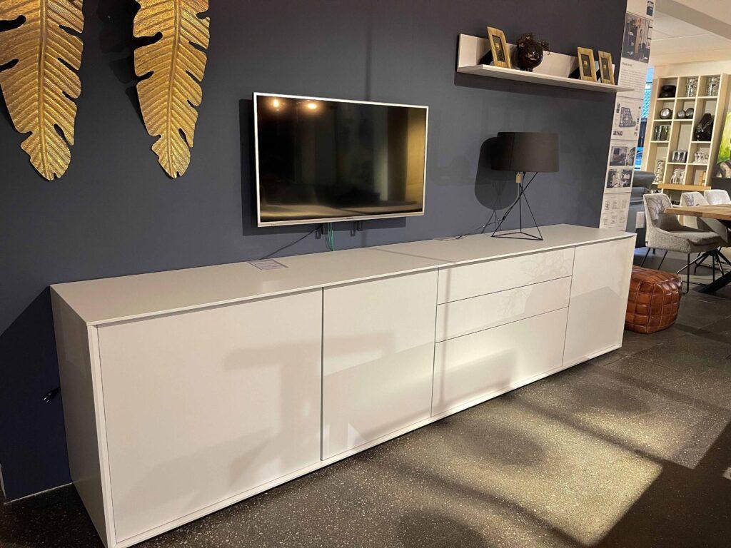 Modernes Wohnzimmer mit dunkelblauer Wand, an der ein flacher Fernseher montiert ist. Darunter steht ein großes, weißes Sideboard mit glatten Fronten und Schubladen, das sich über die gesamte Wandbreite erstreckt. Auf dem Sideboard steht eine moderne schwarze Tischlampe mit dreibeinigem Gestell. Über dem Fernseher hängt ein schlichtes Regal mit goldgerahmten Bildern und Dekoration. Zwei goldene Deko-Blätter schmücken die Wand links neben dem Fernseher.
