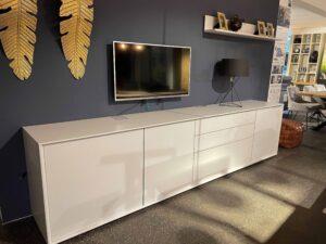 Modernes Wohnzimmer mit dunkelblauer Wand, an der ein flacher Fernseher montiert ist. Darunter steht ein großes, weißes Sideboard mit glatten Fronten und Schubladen, das sich über die gesamte Wandbreite erstreckt. Auf dem Sideboard steht eine moderne schwarze Tischlampe mit dreibeinigem Gestell. Über dem Fernseher hängt ein schlichtes Regal mit goldgerahmten Bildern und Dekoration. Zwei goldene Deko-Blätter schmücken die Wand links neben dem Fernseher.