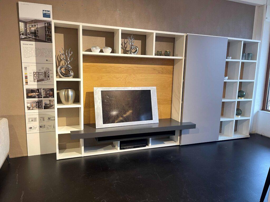 Moderne Wohnwand aus dem Programm „Global 4450“ mit offenen Regalmodulen in Weiß, einer grauen Schiebetür und einem zentralen Wandpaneel in Holzoptik. In der Mitte befindet sich ein graues TV-Board mit einem Fernseher. Die Regale sind mit dekorativen Objekten wie Vasen und Skulpturen bestückt. Links steht eine Infotafel mit Planungsbeispielen und technischen Details.