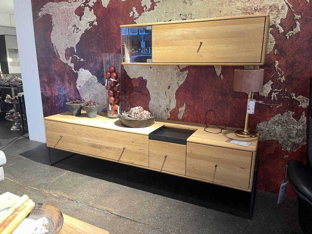 Sideboard und Hängeschrank aus hellem Holz vor einer eindrucksvollen Weltkarten-Tapete in Rot- und Beigetönen. Auf dem Sideboard stehen dekorative Elemente wie Schalen mit Trockenblumen, eine hohe Glasvase mit roten Kugeln und eine Tischleuchte mit goldener Basis und rotem Schirm. Der Hängeschrank ist mittig platziert und ergänzt das minimalistische, moderne Design des Möbelstücks.