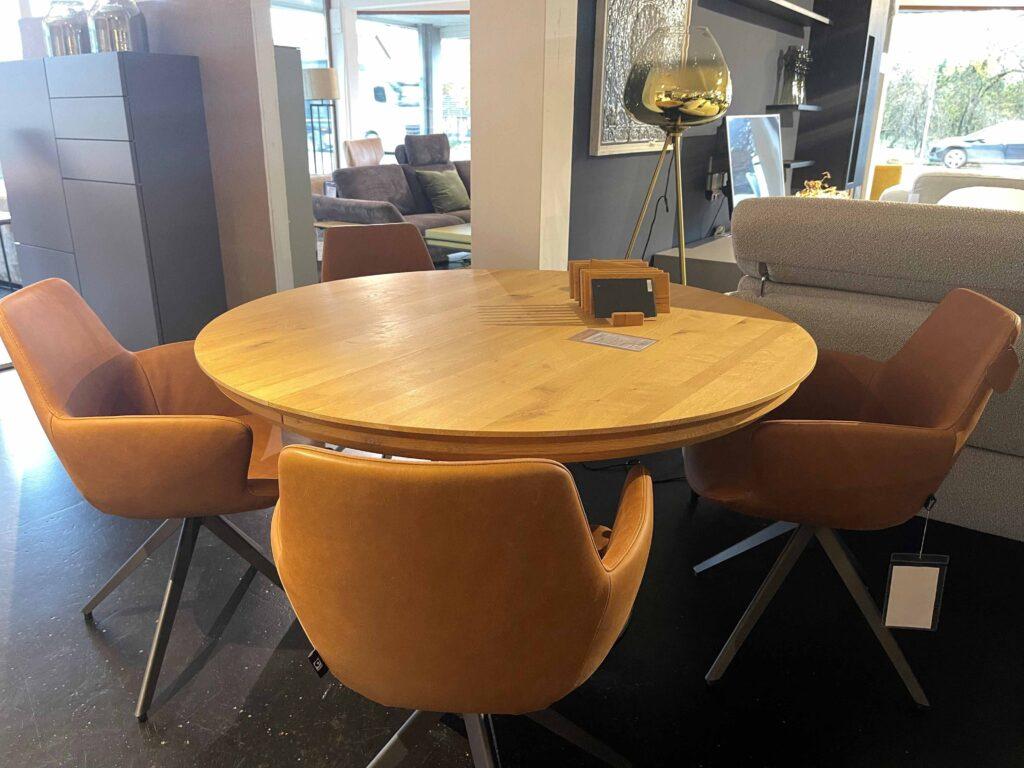 Runder Esstisch aus Holz mit vier gepolsterten Stühlen in warmem Cognac-Ton. Die Stühle haben Armlehnen und ein modernes Kreuzfuß-Gestell aus Metall. Der Tisch steht in einem Ausstellungsraum mit verschiedenen Wohnmöbeln, im Hintergrund sind ein graues Sofa, ein dunkelblauer Wandschrank und eine große Stehlampe mit Glas-Schirm zu sehen.