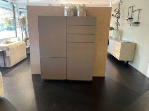 Modernes Sideboard mit klarer Linienführung und matter grauer Oberfläche, bestehend aus Türen und Schubladen. Es steht vor einer halbhohen Raumtrennwand in einem Ausstellungsraum mit dunklem Boden. Auf dem Sideboard befinden sich große Glasvasen. Im Hintergrund sind weitere Möbelstücke und Wohnaccessoires sichtbar.