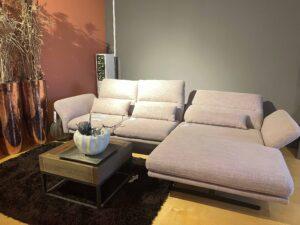 Modernes Wohnzimmer-Arrangement mit einer großen, hellrosa Eckcouch mit verstellbaren Armlehnen. Die Couch steht auf einem hellen Holzboden und ist auf einem dunkelbraunen Hochflor-Teppich platziert. Davor befindet sich ein kleiner quadratischer Couchtisch aus Holz und schwarzem Metallgestell mit einer weißen, kürbisförmigen Dekoschale. Im Hintergrund stehen zwei große glänzende Kupfervasen mit getrocknetem Gräser-Arrangement. Die Wand ist zweifarbig gestrichen – links in Terrakotta, rechts in Grau.