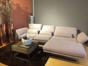 Modernes Wohnzimmer-Arrangement mit einer großen, hellrosa Eckcouch mit verstellbaren Armlehnen. Die Couch steht auf einem hellen Holzboden und ist auf einem dunkelbraunen Hochflor-Teppich platziert. Davor befindet sich ein kleiner quadratischer Couchtisch aus Holz und schwarzem Metallgestell mit einer weißen, kürbisförmigen Dekoschale. Im Hintergrund stehen zwei große glänzende Kupfervasen mit getrocknetem Gräser-Arrangement. Die Wand ist zweifarbig gestrichen – links in Terrakotta, rechts in Grau.