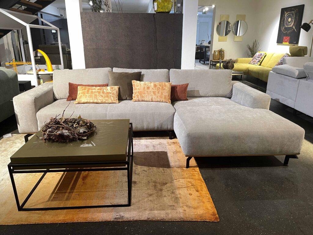 Modernes Wohnzimmer mit einer großen, hellgrauen Eckcouch mit Recamiere auf der rechten Seite. Die Couch ist mit mehreren Dekokissen in Beige-, Braun- und Ockertönen ausgestattet. Davor steht ein quadratischer Couchtisch mit einem olivgrünen Tischblatt und einem schwarzen Metallgestell. Auf dem Tisch liegt eine natürliche Dekoration aus Zweigen. Der Boden ist mit einem Teppich in Beige- und Brauntönen ausgelegt. Im Hintergrund sind weitere Sitzmöbel und Wohnaccessoires sichtbar.