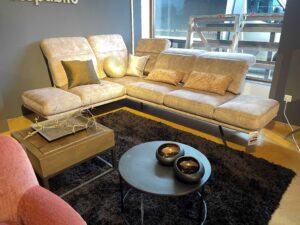 Modern eingerichtetes Wohnzimmer mit einer beigefarbenen Eckcouch aus Mikrofaser mit mehreren dekorativen Kissen in Gold- und Bronzetönen. Die Couch steht auf einem dunkelbraunen Hochflor-Teppich. Vor der Couch befinden sich zwei Couchtische: einer aus Holz mit Metallgestell und dekorativem Ast, und ein runder schwarzer Tisch mit zwei glänzenden, schwarzen Schalen. Im Hintergrund große Fenster mit Blick auf ein Fahrzeug und eine graue Wand mit dem Schriftzug „republic“.