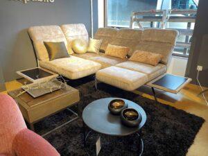 Modern eingerichtetes Wohnzimmer mit einer beigefarbenen Eckcouch aus Mikrofaser mit mehreren dekorativen Kissen in Gold- und Bronzetönen. Die Couch steht auf einem dunkelbraunen Hochflor-Teppich. Vor der Couch befinden sich zwei Couchtische: einer aus Holz mit Metallgestell und dekorativem Ast, und ein runder schwarzer Tisch mit zwei glänzenden, schwarzen Schalen. Im Hintergrund große Fenster mit Blick auf ein Fahrzeug und eine graue Wand mit dem Schriftzug „republic“.