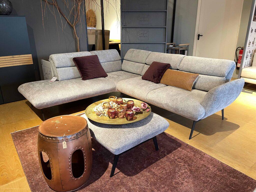 Modernes Wohnzimmer mit einer großzügigen, hellgrauen Eckcouch auf schwarzen, filigranen Beinen. Die Couch ist mit weinroten und ockerfarbenen Kissen dekoriert. Davor steht ein passender Hocker, auf dem ein goldfarbenes Tablett mit dekorativen Gläsern und Trockenblumen platziert ist. Links steht ein kupferfarbener, trommelartiger Beistelltisch. Der Raum ist mit einem weinroten Teppich und hellem Holzfußboden ausgestattet. Im Hintergrund befinden sich dunkle Schränke, ein Dekobaum und eine offene Tür.