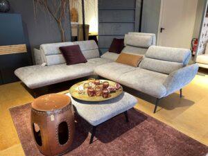 Modernes Wohnzimmer mit einer großzügigen, hellgrauen Eckcouch auf schwarzen, filigranen Beinen. Die Couch ist mit weinroten und ockerfarbenen Kissen dekoriert. Davor steht ein passender Hocker, auf dem ein goldfarbenes Tablett mit dekorativen Gläsern und Trockenblumen platziert ist. Links steht ein kupferfarbener, trommelartiger Beistelltisch. Der Raum ist mit einem weinroten Teppich und hellem Holzfußboden ausgestattet. Im Hintergrund befinden sich dunkle Schränke, ein Dekobaum und eine offene Tür.