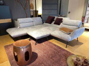 Modernes Wohnzimmer mit einer großzügigen, hellgrauen Eckcouch auf schwarzen, filigranen Beinen. Die Couch ist mit weinroten und ockerfarbenen Kissen dekoriert. Davor steht ein passender Hocker, auf dem ein goldfarbenes Tablett mit dekorativen Gläsern und Trockenblumen platziert ist. Links steht ein kupferfarbener, trommelartiger Beistelltisch. Der Raum ist mit einem weinroten Teppich und hellem Holzfußboden ausgestattet. Im Hintergrund befinden sich dunkle Schränke, ein Dekobaum und eine offene Tür.