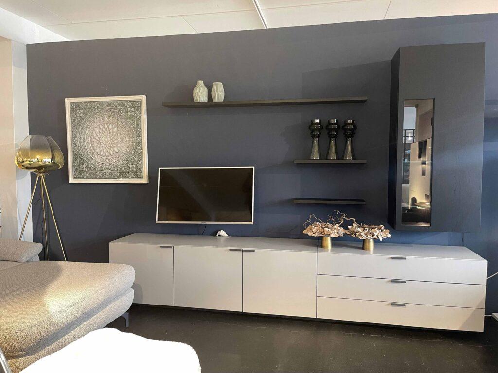Modernes Wohnzimmer mit dunkelblauer Wand, an der eine stilvolle Wohnwand in Hellgrau und Schwarz angebracht ist. Der untere Teil besteht aus geschlossenen Schränken und Schubladen, auf dem ein Fernseher steht. Darüber befinden sich schwebende Wandregale mit dekorativen Vasen und Objekten. Rechts hängt ein schmaler Hängeschrank mit Glaseinsatz. Links steht eine große Stehlampe mit goldfarbenem Glaslampenschirm auf einem Dreibeingestell. Auf der linken Seite ist ein graues Ecksofa angeschnitten sichtbar.