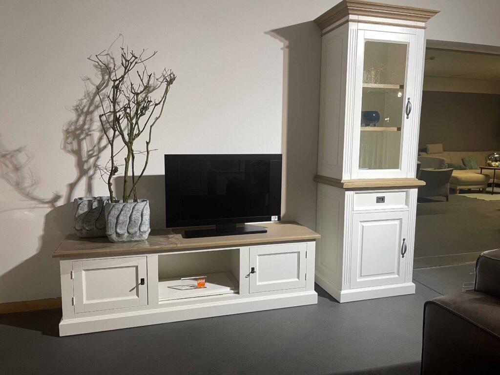 Wohnwand im modernen Landhausstil mit weißem TV-Unterschrank und hohem Vitrinenschrank. Der TV-Schrank besitzt zwei Türen und ein offenes Fach in der Mitte, darauf stehen ein Flachbildfernseher sowie zwei strukturierte, graue Pflanzgefäße mit dekorativen Ästen. Der Vitrinenschrank rechts hat ein Glaselement mit zwei beleuchteten Böden, eine Schublade und eine weitere Tür im unteren Bereich. Die Möbel haben eine helle Holzplatte als Kontrast zur weißen Lackierung.