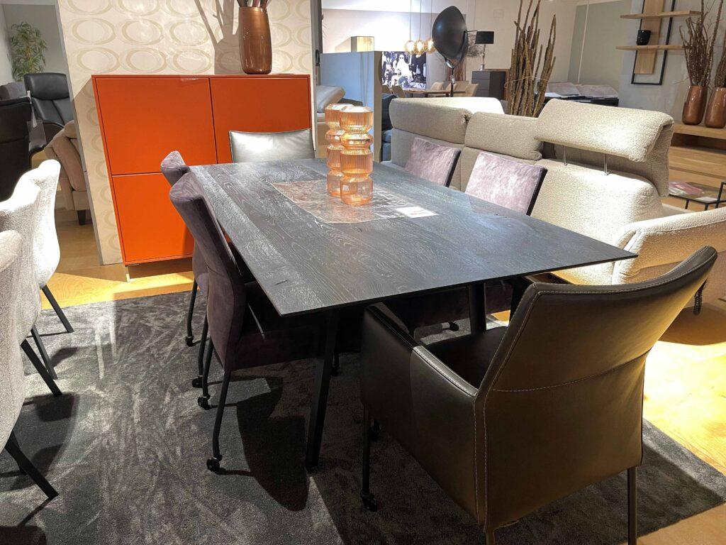 Essgruppe mit modernem, rechteckigem Esstisch aus dunklem Holz mit leicht abgeschrägten Kanten. Rund um den Tisch stehen verschiedene Polsterstühle in Grau- und Schwarztönen, einige mit Armlehnen, andere mit Rollen. Im Hintergrund steht eine auffällige orange Hochkommode mit vier quadratischen Fronten. Auf dem Tisch befinden sich mehrere stapelbare Gläser in warmen Bernsteinfarben als Dekoration. Die Szene ist auf einem dunklen Teppich arrangiert, daneben ein helles Sofa mit verstellbaren Kopfstützen.