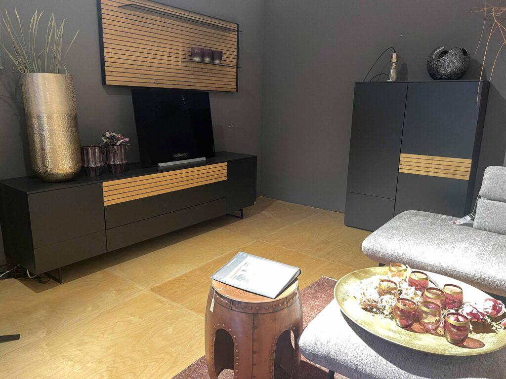 Modernes Wohnzimmer mit dunklen, mattschwarzen Möbeln mit horizontalen Holzlamellen-Akzenten. Links steht ein breites Sideboard mit goldener Vase, Dekoblumen und einem Fernseher von Hülsta. Rechts daneben ein hoher, passender Schrank. An der Wand darüber befindet sich ein Wandpaneel mit Regal und Dekotassen. Im Vordergrund eine graue Polstergarnitur mit einem goldenen Tablett, darauf dekorative Gläser und Trockenblumen. Der Raum hat einen hellen Holzfußboden und eine dunkle Wandfarbe.