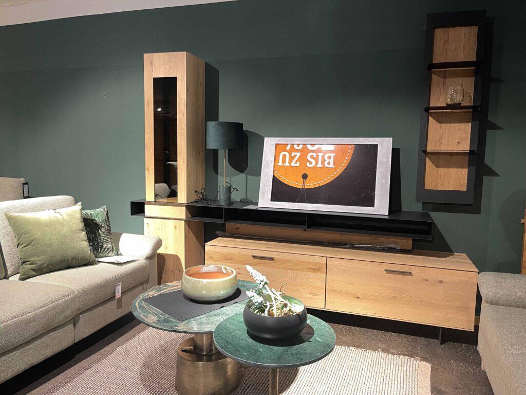Wohnzimmer-Ensemble mit grün gestrichener Wand und modernem Möbel-Set aus Holz und schwarzem Metall. Links steht ein beiger Zweisitzer mit grünen Kissen. Im Zentrum eine TV-Wand mit Sideboard, Hochschrank und Wandregal in Eiche-Optik mit schwarzen Akzenten. Ein Fernseher mit Angebotsplakat „Bis zu …“ lehnt auf dem Sideboard. Davor ein runder Couchtisch mit grünem Marmor-Look und messingfarbenem Sockel, dekoriert mit zwei stilvollen Keramikschalen. Auf dem Boden liegt ein grob gewebter Teppich in Naturtönen.
